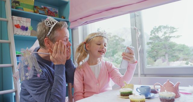 Grandfather and granddaughter wearing tiaras engage in an imaginative tea party at home, creating lasting memories. Great for themes of family bonding, childhood, grandparents, playtime, and joyful moments. Ideal for articles, blogs, advertisements, or cards focused on intergenerational relationships and imaginative play.