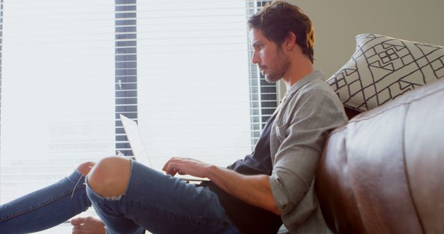 Young man sitting on couch by window, working on laptop. Casual setting with relaxed atmosphere, making this suitable for illustrations of remote work, home office setups, technology use, or lifestyles of young professionals.