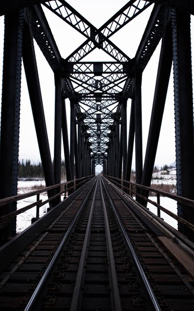 Steel railroad bridge spans over snowy landscape, emphasizing symmetrical perspective. Ideal for use in themes relating to travel, transportation infrastructure, industrial architecture, and winter settings. Suitable for presentations, blogs, and educational materials focusing on engineering and construction.