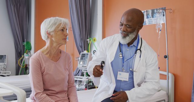 Doctor consulting with senior woman in hospital room. The doctor is explaining something while holding a medical device. The hospital room is equipped with medical equipment in the background. The scene conveys a sense of trust, care, and professional medical service. This stock photo is useful for articles or promotions about healthcare services, elder care, and medical professions.