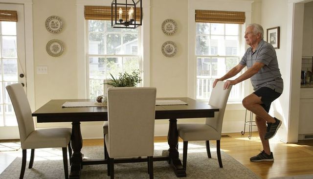 Senior man balancing on one leg while holding chair in sunlit modern home dining room