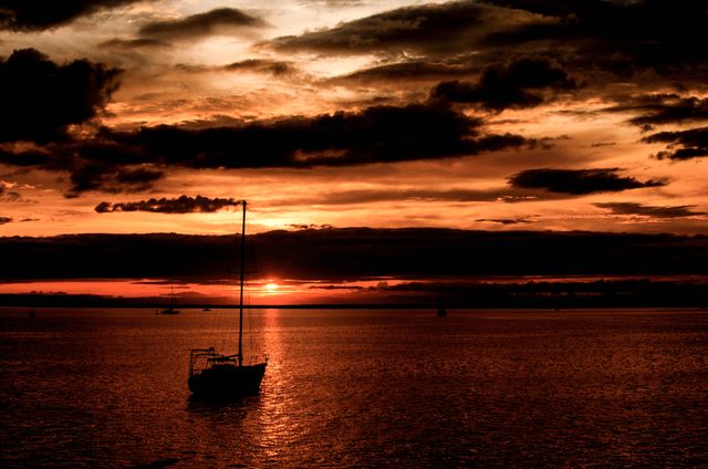 This stunning image captures a sailboat silhouetted against a dramatic sunset over a tranquil sea, with dark clouds adding to the dramatic effect. The scene invokes a sense of peace and adventure, making it perfect for travel brochures, outdoor adventure promotions, relaxation concepts, and inspirational posters.
