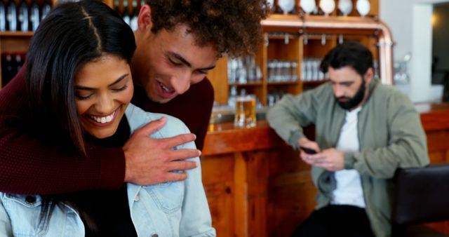 Young interracial couple laughing and embracing in cozy bar while another man appears distracted in the background with his phone. Perfect for themes revolving around love, relationships, bar atmosphere, social interactions, and modern lifestyle scenarios.