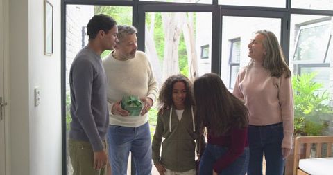 Multigenerational Diverse Family Welcoming at Sunlit Glass Doorway Holding Wrapped Gift