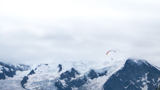 Paraglider soaring over snow-covered mountain peaks with cloudy sky above. Captures thrill and freedom of extreme sports, perfect for travel brochures, adventure sports promotions, or websites related to outdoor activities. Also useful for inspirational themes, emphasizing freedom, exploration, and natural beauty.