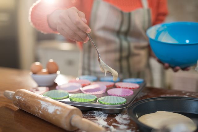 Woman in kitchen wearing apron, spooning muffin batter into colorful paper cases in baking tray. Rolling pin and eggs on wooden table. Ideal for content related to home baking, cooking tutorials, culinary hobbies, and homemade desserts.