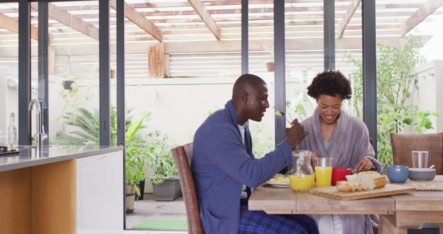An African American couple is enjoying breakfast together at a dining table in a modern, bright indoor space filled with natural light. The scene depicts happiness, bonding, and a healthy lifestyle. This image can be used for promoting family bonding, healthy living, home and kitchen products, or articles related to relationships and daily routines.