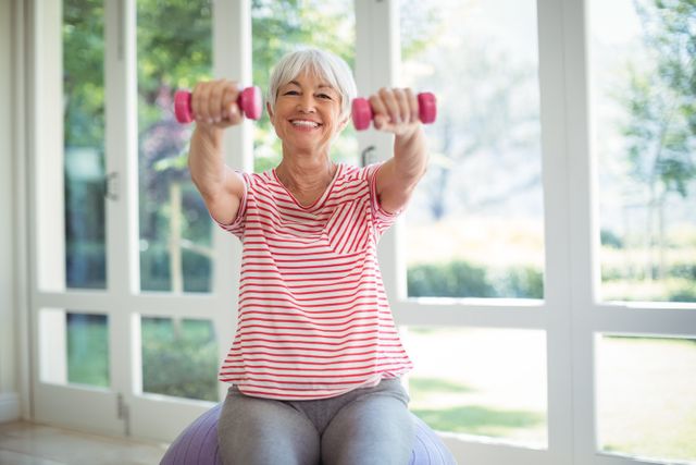 Senior woman sitting on exercise ball, holding pink dumbbells, smiling while working out at home. Bright natural light from large windows creates a cheerful atmosphere. Ideal for promoting active aging, home fitness routines, and healthy lifestyle for seniors.