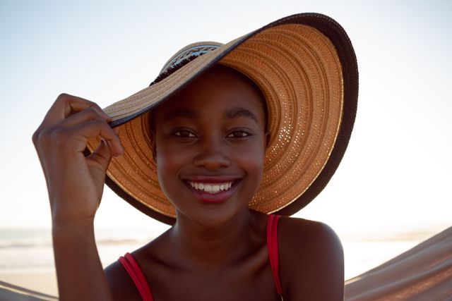 Young woman enjoying a sunny day at the beach while relaxing in a hammock. She is wearing a wide-brimmed sun hat and a red swimsuit, smiling at the camera. Ideal for use in travel brochures, summer vacation promotions, lifestyle blogs, and advertisements focused on relaxation and leisure.