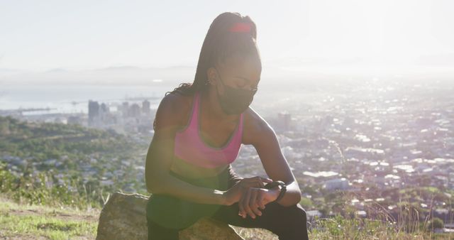 African american woman wearing face mask using smartwatch while sitting on a rock outdoors. fitness sports and active lifestyle during coronavirus pandemic concept