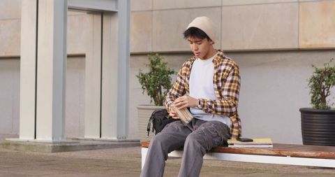 Asian college student checking wallet on campus bench in modern urban courtyard with backpack