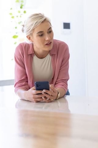 Young woman leaning on glossy kitchen counter holding smartphone in bright modern home