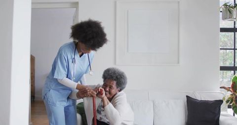 African american nurse helping elderly woman with cane on living room sofa, home care support