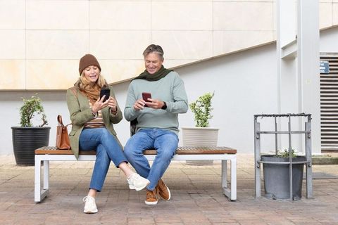 Senior couple sitting on bench scrolling smartphones in urban plaza, smiling together