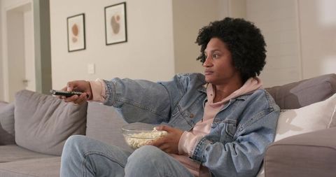African american man lounging on sofa watching tv with remote and popcorn, cozy evening
