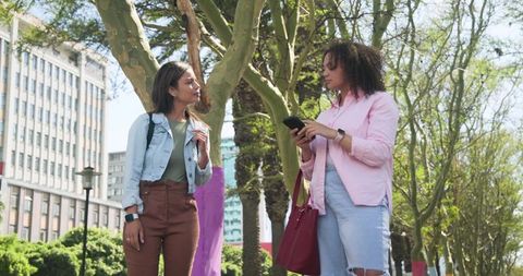 Diverse women standing and talking on sunny city walkway holding smartphone and red tote