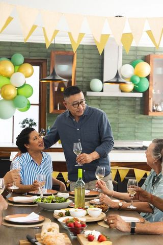 Senior asian man raising toast at diverse family dinner party with balloons and garland