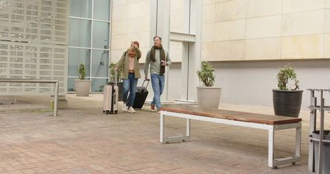 Senior couple walking with luggage and drinking coffee in urban courtyard