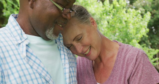 This stock photo features an interracial senior couple sharing an intimate and loving moment outdoors. The couple is smiling and showing affection, with greenery in the background suggesting a peaceful nature setting. Perfect for use in topics related to senior lifestyles, diversity in relationships, retirement happiness, and romance in older age.