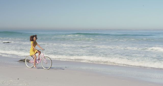 Happy biracial woman riding bicycle on sunny beach by the sea, with copy space. Summer, free time, relaxation and vacations.