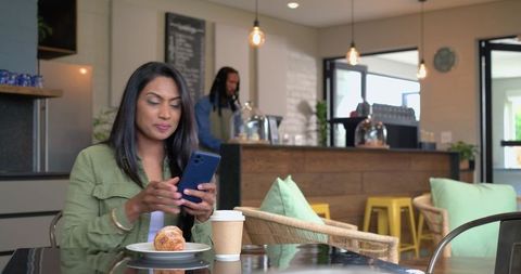 Woman scrolling phone while enjoying croissant and coffee in cozy urban café