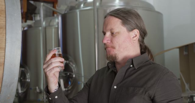 Craft brewer closely inspecting a glass of beer in a brewery, surrounded by industrial stainless steel tanks. Man is wearing a dark shirt, carefully evaluating the beer's color and clarity. Ideal for depicting quality control in the craft beer industry, marketing campaigns for breweries, beverage industry visuals, and professional craftsmanship promotions.