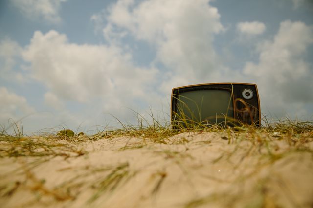 Retro television lying in untouched sandy beach with a cloudy sky in the background, creating a sense of nostalgia and obsolescence. Suitable for illustrating contrast between old technology and natural landscapes, and concepts related to technology abandonment, time passing, and unpredictable encounters in unusual places. Ideal for creative projects, blogs, articles, and advertisements focusing on environmental awareness, outdated tech, or artistic expression.