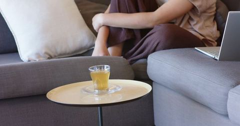 Woman sitting hugging knees on grey sofa with glass of tea and laptop, cozy remote work