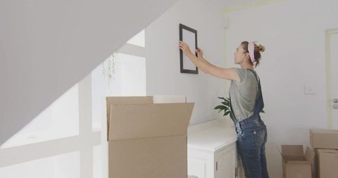 Mid-adult woman hanging frame above sideboard while unpacking in bright minimalist living space