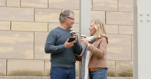 Mature couple enjoying coffee and friendly conversation on urban plaza near stone wall
