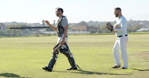 Male baseball teammates walking across sunny field carrying catchers gear