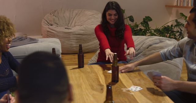 High angle view of a group of five young multi-ethnic male and female friends sitting around a table playing cards and making a toast with bottles of beer in the sitting room of an apartment at night