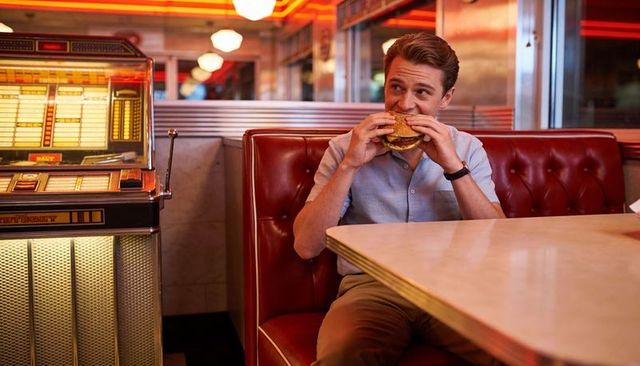 Man enjoying burger in neon retro diner booth with vintage jukebox and warm reflections