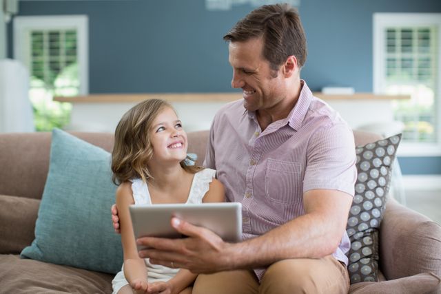 Smiling father and daughter sitting on sofa using digital tablet in living room at home