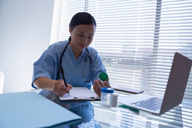 Female doctor in blue scrubs writing a prescription on a clipboard while sitting at a desk in a hospital clinic. She has a stethoscope around her neck and is using a laptop. Ideal for use in healthcare, medical, and hospital-related content, illustrating professional medical practice and patient care.
