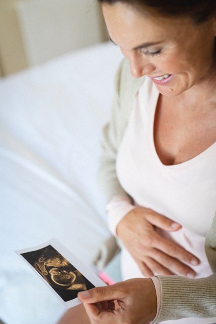 Close-up of smiling caucasian mid adult pregnant woman holding ultrasound scan report. unaltered, maternity, anticipation, beginnings, prenatal care, copy space, medical scan and domestic life.