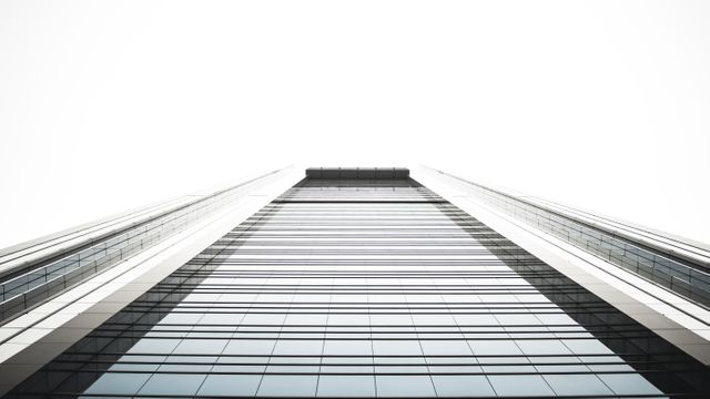 Described image displays a low-angle view of a glass skyscraper against a white sky. Shows architectural details of the modern building and conveys impressions of height, strategic design, and urban sophistication. Great for use in corporate brochures, presentations, real estate advertisements, urban development articles, and stock photos illustrating professional environments.