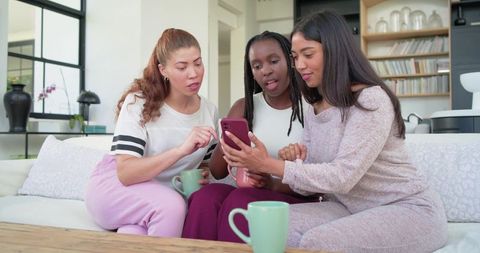 Three friends relaxing on couch sharing smartphone and coffee in cozy modern living room