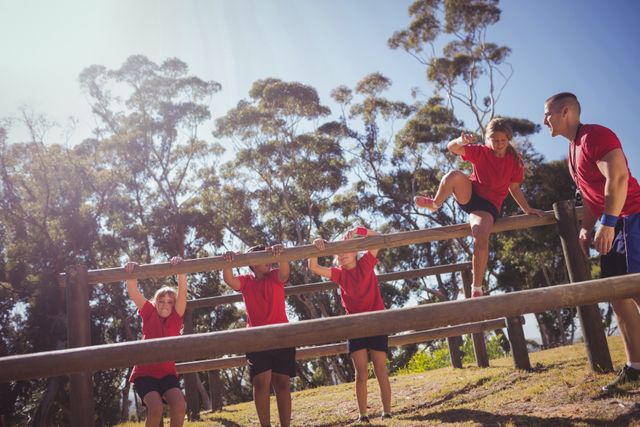 Trainer guiding kids through an obstacle course in a boot camp. Children wearing red shirts actively participating in outdoor fitness training. Ideal for use in articles about physical education, teamwork, children's fitness programs, and summer camp activities.