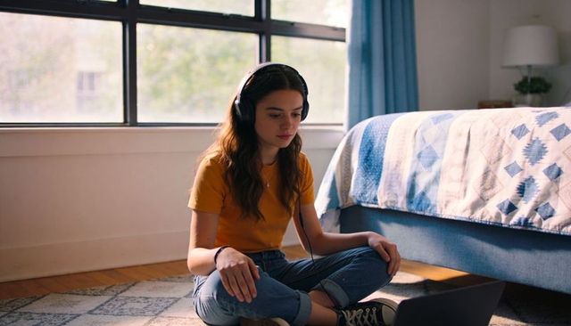 Teen girl focusing on laptop while wearing headphones and sitting on bedroom floor