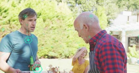 Senior man serving grilled food at backyard gathering, guest holding bottle on sunny patio