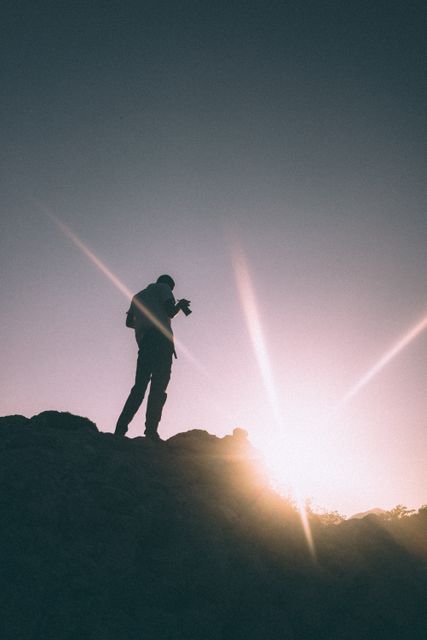 Silhouette of a photographer capturing the sunrise atop a mountain peak. The image highlights the interplay of light flare and the morning light, reflecting a dynamic and adventurous spirit. Ideal for use in travel, photography, and nature-related content, as well as advertisements promoting outdoor activities, adventure travel, and inspirational visuals.