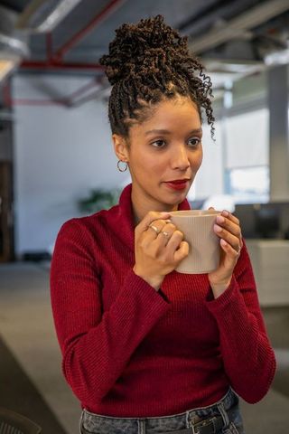 African american woman sipping coffee in modern open-plan office wearing red sweater