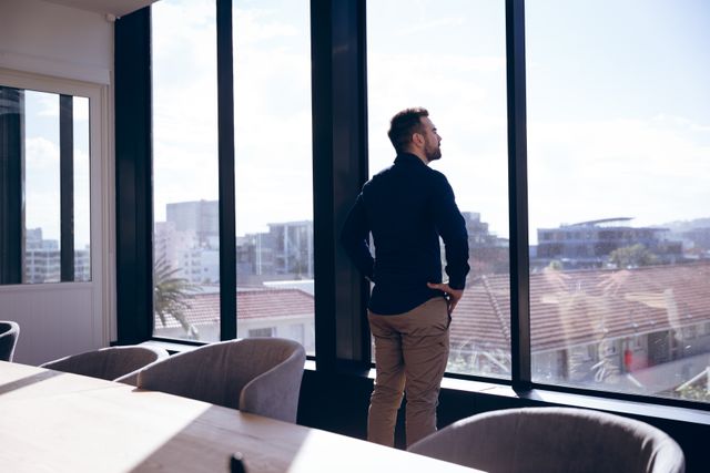 Businessman taking a break and looking out the window in a modern office. Ideal for use in business, corporate, and professional settings, as well as articles on work-life balance, office environments, and urban living.