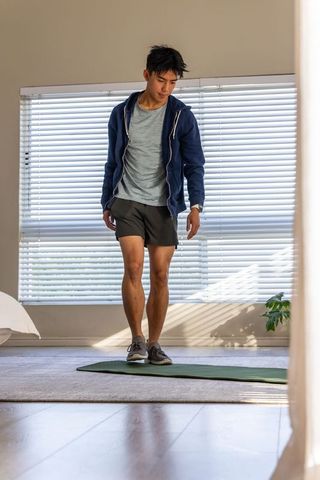 Asian man stretching on green yoga mat in sunlit bedroom wearing navy hoodie