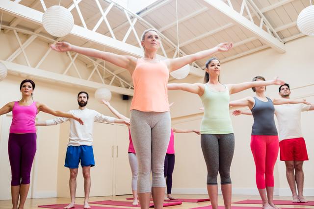 Diverse group of men and women performing stretching exercises in a bright, spacious fitness studio with high ceilings and white walls. Ideal for promoting fitness classes, wellness programs, active and healthy lifestyles, yoga sessions, and gym memberships.