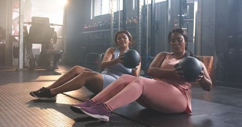 Diverse women doing russian twists with medicine balls in gym, strength training