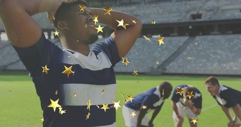 Holding hands-behind-head rugby player on stadium turf with teammates and gold stars
