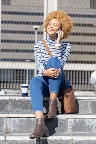 Sunlit african american woman sitting on steps talking on phone with coffee and bag