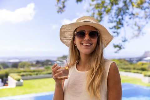 Smiling woman wearing straw sun hat and sunglasses holding lemon water by backyard pool